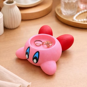 Puede incluir: Un soporte para anillos rosa con forma de personaje de dibujos animados, con alas rojas y ojos azules. El soporte, que contiene dos anillos, está sobre una superficie de madera. El fondo incluye un espejo, frascos de perfume y un pequeño jarrón.
