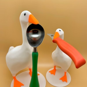 May include: Two white goose figurines with orange beaks and feet. One goose is holding a green handled spoon, the other is holding a red handled knife.