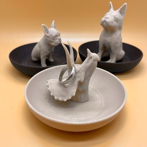 May include: Three white ceramic ring dishes, each featuring a different animal design. One dish has a triceratops, another has a French bulldog, and the third has a French bulldog. A silver ring is resting on the triceratops dish.