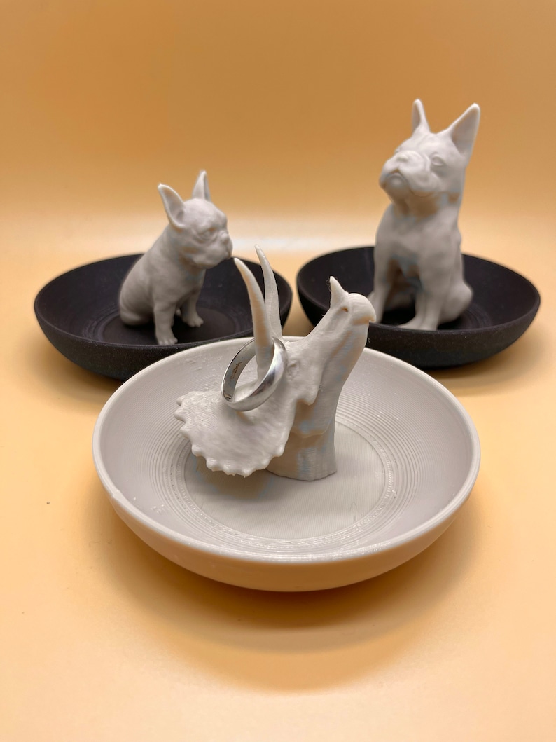 May include: Three white ceramic ring dishes, each featuring a different animal design. One dish has a triceratops, another has a French bulldog, and the third has a French bulldog. A silver ring is resting on the triceratops dish.