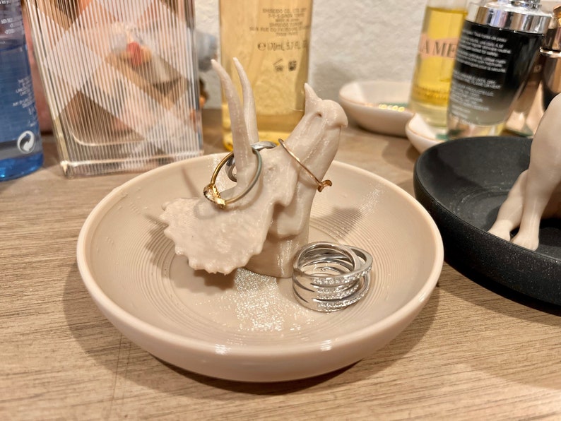 May include: A beige ceramic ring dish shaped like a dinosaur with a textured surface. The dish has a gold ring and a stack of silver rings on it.