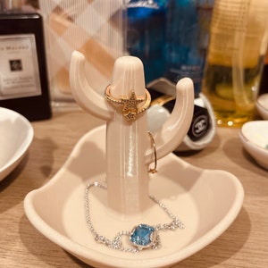 K&ouml;nnte beinhalten: Wei&szlig;er Keramik-Kaktus-Ringst&auml;nder mit einem sternf&ouml;rmigen Ring und einem blauen Edelstein-Armband.