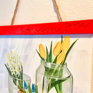 May include: A red wooden hanging sign with the text "i love you" in white lettering. The sign is hanging from a string and is in front of a painting of flowers in vases.