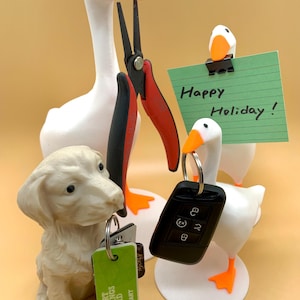 May include: A white goose figurine holding a pair of red pliers, a smaller white goose figurine holding a green note with the text "Happy Holiday!" and a white dog figurine holding a set of keys and a green card with the text "SMART SAVINGS CARD H MART".