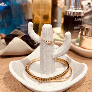 K&ouml;nnte beinhalten: Wei&szlig;er Keramik-Kaktus-Ringst&auml;nder mit zwei goldenen Ringen. Der obere Ring hat eine Herzform. Der untere Ring hat die Worte "Live Laugh Love" eingraviert.