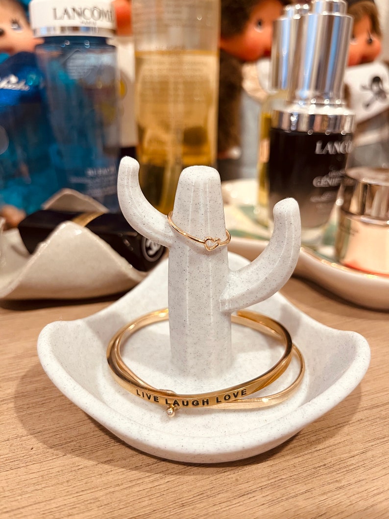 K&ouml;nnte beinhalten: Wei&szlig;er Keramik-Kaktus-Ringst&auml;nder mit zwei goldenen Ringen. Der obere Ring hat eine Herzform. Der untere Ring hat die Worte "Live Laugh Love" eingraviert.