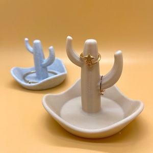 K&ouml;nnte beinhalten: Zwei Keramik-Kaktus-Ringhalter. Einer ist hellblau und der andere beige. Beide haben eine kleine Schale an der Basis und einen Ringhalter in der Mitte. Der beige Kaktus hat einen goldenen Ring mit einem Sternmuster.