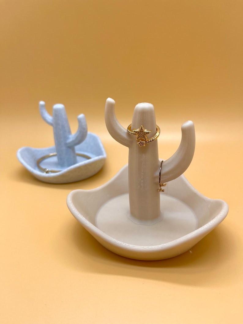 K&ouml;nnte beinhalten: Zwei Keramik-Kaktus-Ringhalter. Einer ist hellblau und der andere beige. Beide haben eine kleine Schale an der Basis und einen Ringhalter in der Mitte. Der beige Kaktus hat einen goldenen Ring mit einem Sternmuster.