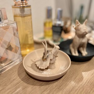 May include: A beige ceramic ring holder shaped like a dinosaur with two gold rings on top. The ring holder is sitting on a beige glitter-covered dish.