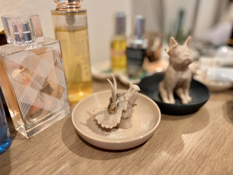 May include: A beige ceramic ring holder shaped like a dinosaur with two gold rings on top. The ring holder is sitting on a beige glitter-covered dish.