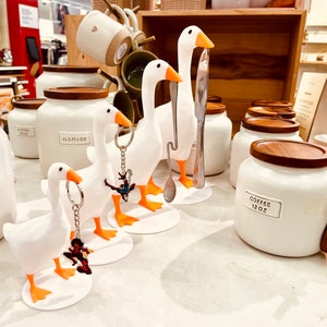 May include: White ceramic canisters with wooden lids and labels that read "Coffee 12 oz" and "All Purpose 48 oz".  Three white goose-shaped ceramic utensil holders with orange feet and keychains attached.
