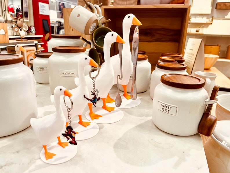 May include: White ceramic canisters with wooden lids and labels that read "Coffee 12 oz" and "All Purpose 48 oz".  Three white goose-shaped ceramic utensil holders with orange feet and keychains attached.