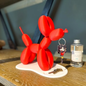 Peut inclure: Une sculpture de chien ballon rouge vif sur une base blanche, avec un porte-clés Mickey Mouse et un petit flacon de parfum. L'ensemble est un objet décoratif fantaisiste, idéal pour un bureau ou une étagère.