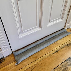 May include: A grey fabric door draught stopper with a white zipper. The draught stopper is placed at the bottom of a white door with a wood frame.