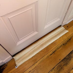 May include: A beige fabric door draft stopper placed at the base of a light pink door. The door has rectangular panels. The stopper is on a wooden floor.