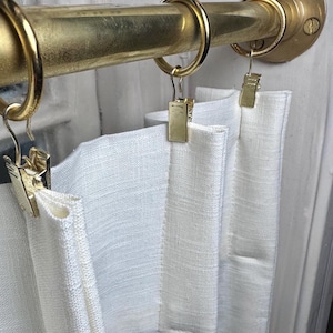 May include: A gold-toned metal curtain rod mounted with a circular wall bracket, featuring three matching gold rings. Each ring is equipped with a gold metal clip that securely holds a white, textured fabric curtain panel in place.