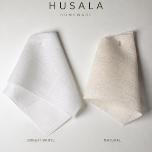 Op de afbeelding: Twee gevouwen stofstalen met het label HUSALA HOMEWARE. Het linker monster is helderwit en het rechter monster heeft een lichte natuurlijke beige kleur. De tekst onderaan luidt: Two small, folded fabric samples are shown against a white background. The fabric on the left is bright white, while the one on the right is a light natural colour.
