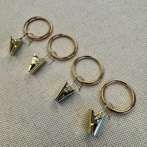 May include: Four gold-colored curtain rings with attached clips. The rings are circular and have a smooth finish. The clips are rectangular with a spring-loaded mechanism for holding fabric. The items are arranged diagonally on a textured, light-colored surface.