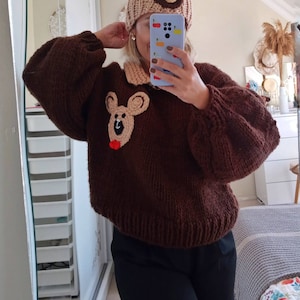 Può includere: Un maglione di lana marrone con un'applicazione a forma di orso marrone sul davanti. Il maglione ha un colletto bianco e un cappello a forma di orso marrone con un pon pon marrone in cima.