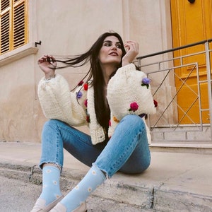 Puede incluir: Una mujer lleva un c&aacute;rdigan blanco de punto con bordados florales de colores, vaqueros azules y calcetines azul claro con estampados de margaritas blancas. Lleva mocasines blancos y est&aacute; sentada en una acera.