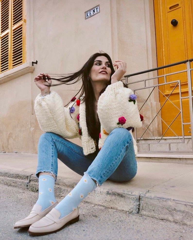 Puede incluir: Una mujer lleva un c&aacute;rdigan blanco de punto con bordados florales de colores, vaqueros azules y calcetines azul claro con estampados de margaritas blancas. Lleva mocasines blancos y est&aacute; sentada en una acera.