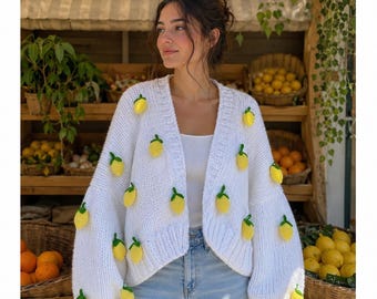 Può includere: Un cardigan in maglia bianca con scollo a V e maniche lunghe, decorato con molteplici applicazioni tridimensionali a forma di limone giallo con foglie verdi. Il capo è indossato sopra un top bianco e jeans azzurri.