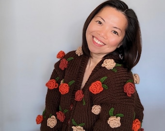 Può includere: Un cardigan in maglia marrone scuro con scollo a V e chiusura frontale con bottoni. Il capo è decorato con applicazioni floreali all'uncinetto tridimensionali nei toni del crema, arancione e rosso, ciascuna accentuata da piccole foglie verdi.