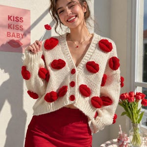 May include: A cream-colored knit cardigan featuring a V-neckline, long sleeves, and a button-front closure. The garment is decorated with multiple three-dimensional red lip-shaped appliqués scattered across the front and sleeves.