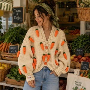 May include: A cream-colored, chunky knit V-neck cardigan featuring three-dimensional orange carrot appliqués with green tops scattered across the front and sleeves. The garment has a ribbed hem and cuffs, and is fastened with three round orange buttons.