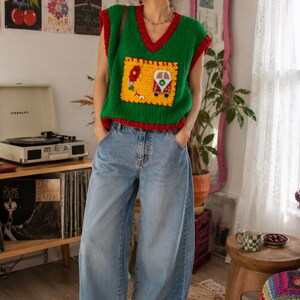 May include: A green sleeveless knit sweater vest with a deep V-neck and red trim, featuring a large yellow square patch on the front with a crocheted van and balloon design, paired with light blue wide-leg denim jeans and red canvas sneakers.