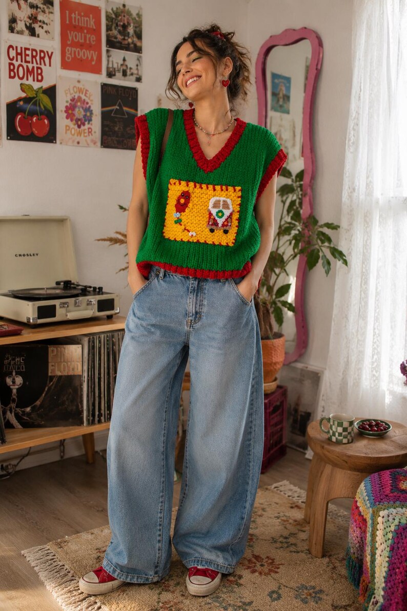 May include: A green sleeveless knit sweater vest with a deep V-neck and red trim, featuring a large yellow square patch on the front with a crocheted van and balloon design, paired with light blue wide-leg denim jeans and red canvas sneakers.
