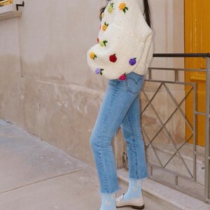 Puede incluir: Un su&eacute;ter de crochet blanco con flores bordadas de colores. El su&eacute;ter se lleva con jeans azules y calcetines azul claro con patrones de margaritas blancas. La persona lleva mocasines blancos.