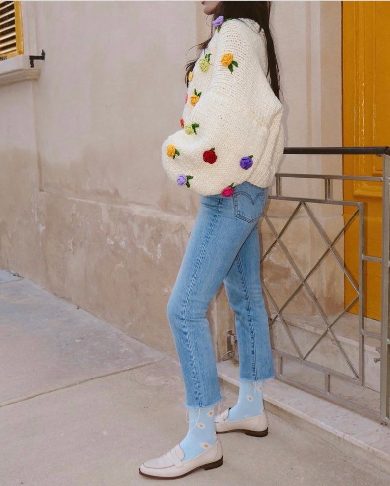Puede incluir: Un su&eacute;ter de crochet blanco con flores bordadas de colores. El su&eacute;ter se lleva con jeans azules y calcetines azul claro con patrones de margaritas blancas. La persona lleva mocasines blancos.