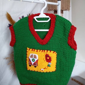 May include: A green sleeveless knit sweater vest with a V-neckline and red trim around the neck, armholes, and hem. A large yellow rectangular patch is stitched to the front, featuring a small crocheted van and a red kite with a long tail.