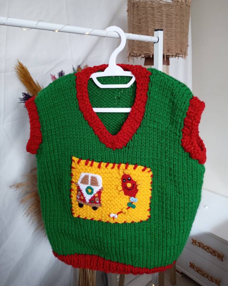 May include: A green sleeveless knit sweater vest with a V-neckline and red trim around the neck, armholes, and hem. A large yellow rectangular patch is stitched to the front, featuring a small crocheted van and a red kite with a long tail.