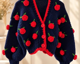 May include: A navy blue knit cardigan featuring a bright red trim along the V-neckline, cuffs, and hem. The garment is decorated with multiple three-dimensional red rose appliqués, each accented with small green crocheted leaves.