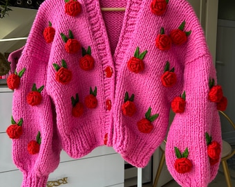 May include: A pink knitted cardigan with a V-neck and button closure. The cardigan is decorated with red and green 3D flower embellishments.