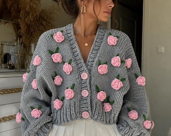 Può includere: Un cardigan in maglia grossa grigia con scollo a V, maniche lunghe con polsini a coste e tre grandi bottoni rosa chiaro. Il capo è decorato con numerose rose all'uncinetto tridimensionali di colore rosa chiaro con piccole foglie verdi.