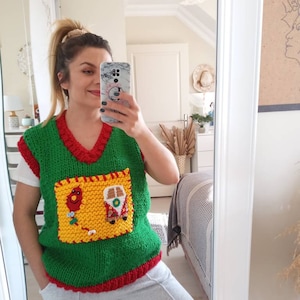 May include: A green knitted sleeveless sweater vest with a red V-neck collar and hem. It features a large, square yellow patch pocket on the front, decorated with a small red balloon and a colorful camper van applique with intricate details.