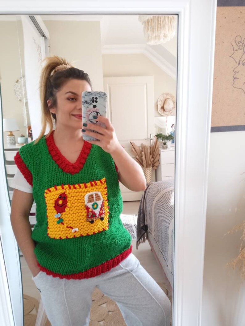May include: A green knitted sleeveless sweater vest with a red V-neck collar and hem. It features a large, square yellow patch pocket on the front, decorated with a small red balloon and a colorful camper van applique with intricate details.