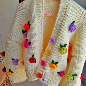 Puede incluir: Un c&aacute;rdigan de punto amarillo con adornos de flores de colores y cierres de botones. El c&aacute;rdigan tiene cuello en V y mangas largas.
