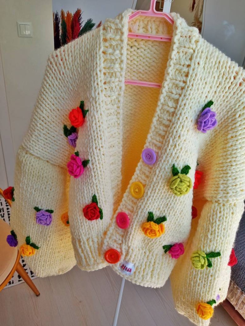 Puede incluir: Un c&aacute;rdigan de punto amarillo con adornos de flores de colores y cierres de botones. El c&aacute;rdigan tiene cuello en V y mangas largas.