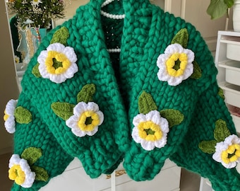 May include: A chunky knit cardigan in a vibrant emerald green color, featuring large, three-dimensional crocheted flower appliqués. Each flower has white petals, a bright yellow center, and green leaves attached to the thick, textured yarn.