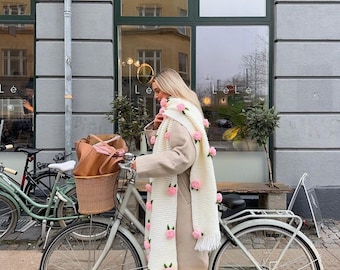 May include: A woman in a beige coat and blue jeans is standing next to a white bicycle with a brown wicker basket. She is wearing a white scarf with pink flower embellishments. The bicycle is parked in front of a building with a large window.