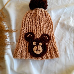 Könnte beinhalten: Eine gestrickte braune Mütze mit einem braunen Bommel oben und einem gestickten braunen Bärenkopf auf der Vorderseite.