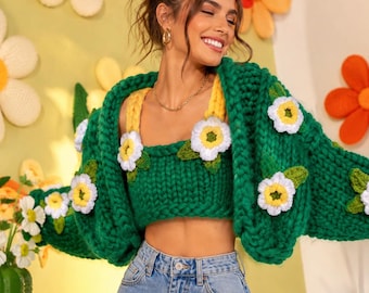 May include: A chunky knit emerald green cardigan and matching crop top set featuring three-dimensional white and yellow crocheted flower appliqués with green leaves, paired with light blue denim jeans with a classic button and zipper fly closure.