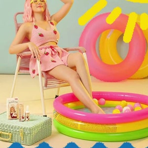 Pode incluir: Um top de croch&ecirc; rosa e saia combinando com estampa de morangos vermelhos. O conjunto &eacute; usado com &oacute;culos de arma&ccedil;&atilde;o amarela e cabelo rosa. Perto est&atilde;o uma mala de vime verde, um frasco de Skinlucent e uma piscina infl&aacute;vel.