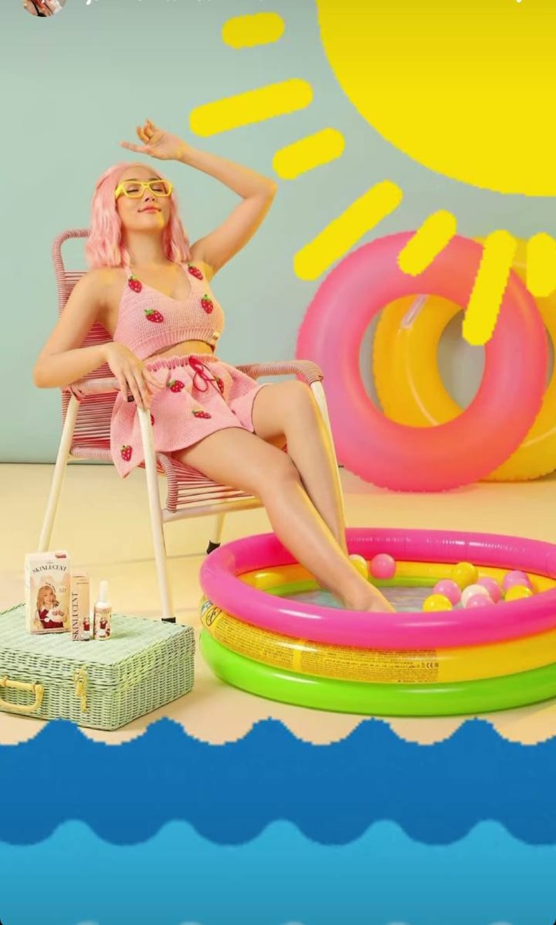 Pode incluir: Um top de croch&ecirc; rosa e saia combinando com estampa de morangos vermelhos. O conjunto &eacute; usado com &oacute;culos de arma&ccedil;&atilde;o amarela e cabelo rosa. Perto est&atilde;o uma mala de vime verde, um frasco de Skinlucent e uma piscina infl&aacute;vel.