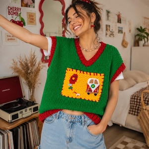 May include: A green knit sweater vest with a red V-neck trim and armholes. The front features a large yellow rectangular patch with a red balloon graphic and a colorful van illustration. The vest is worn over a white short-sleeve t-shirt and blue jeans.