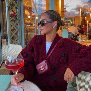 May include: A woman wearing a maroon knitted cardigan with 3D rose embellishments, black sunglasses, and a small burgundy crossbody bag. She is sitting at a table with a glass of wine.